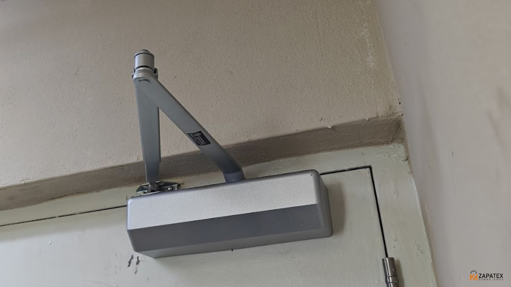 Door closer plateado instalado en la parte superior de una puerta comercial blanca