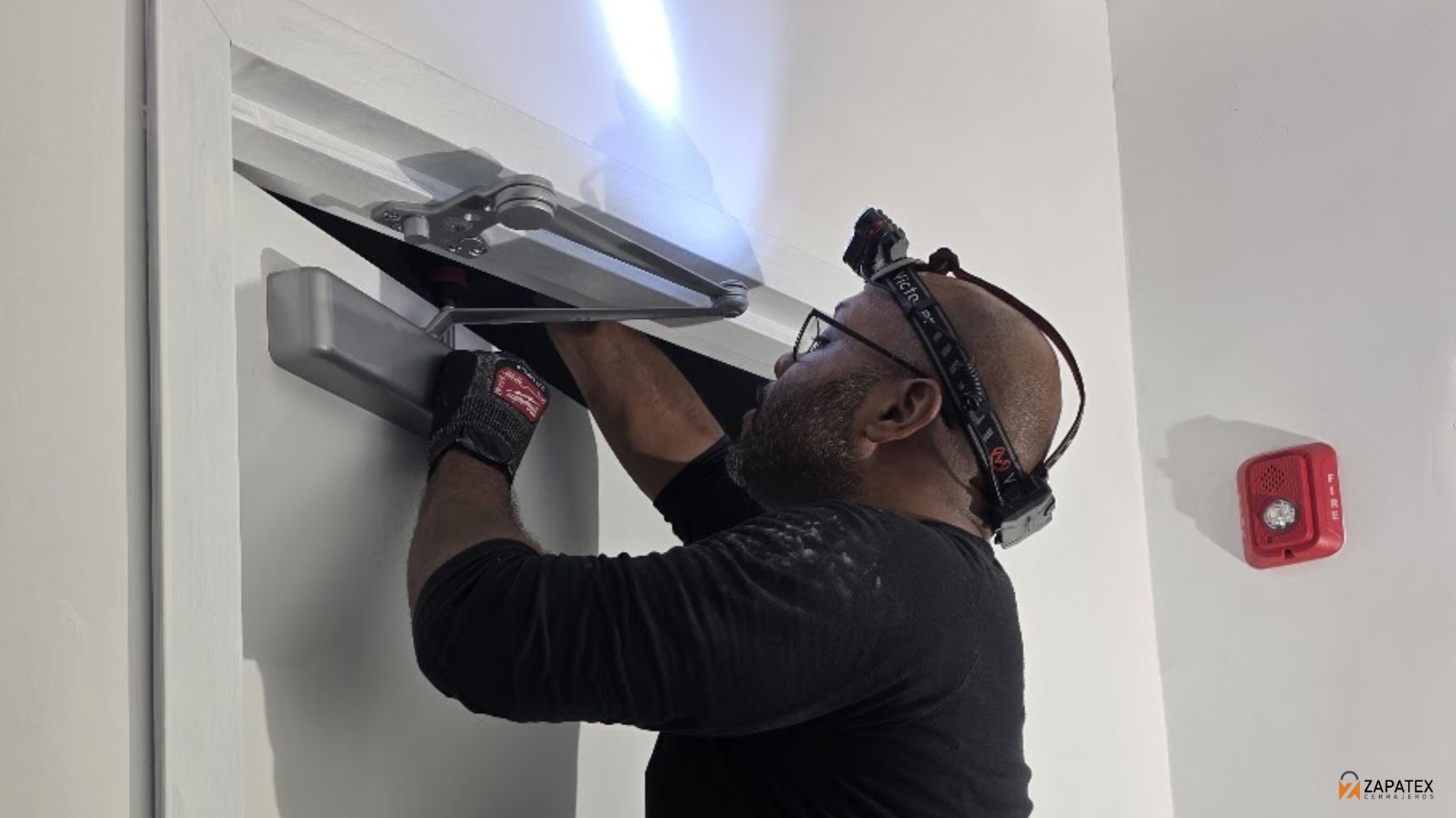 Tecnico colocando una pieza plateada en la parte superior de una puerta junto a un sensor de alarma rojo en la pared